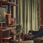 Man seated in a sunlit living room reading a book while a desktop monitor on a wooden desk shows a music player interface nearby.
