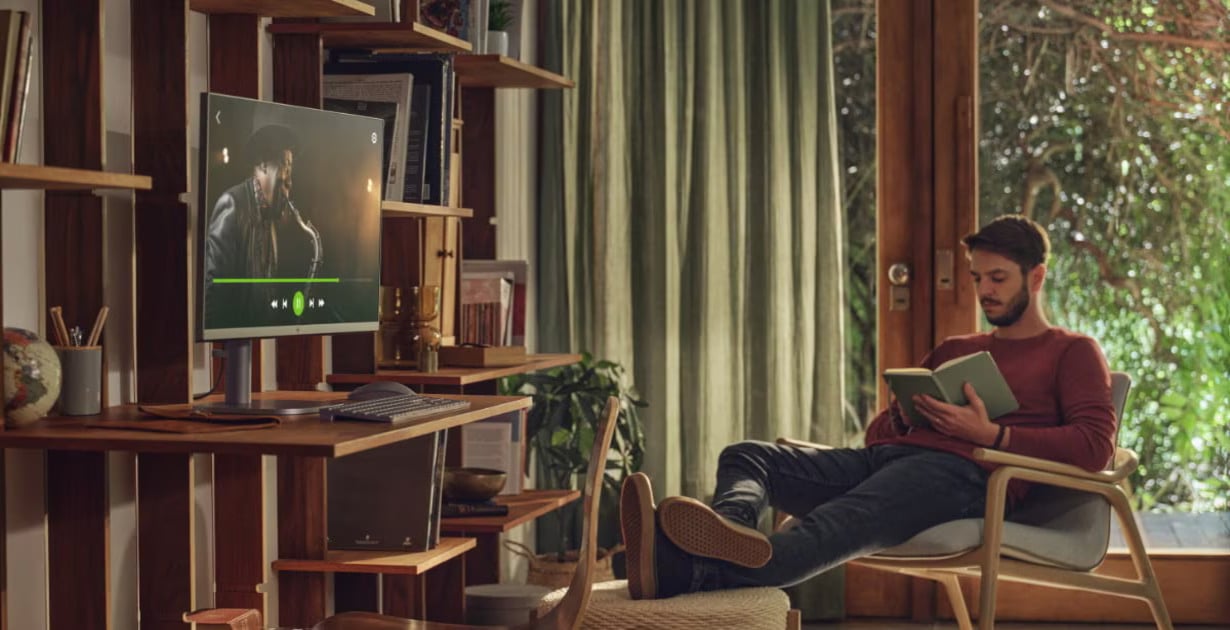 Man seated in a sunlit living room reading a book while a desktop monitor on a wooden desk shows a music player interface nearby.