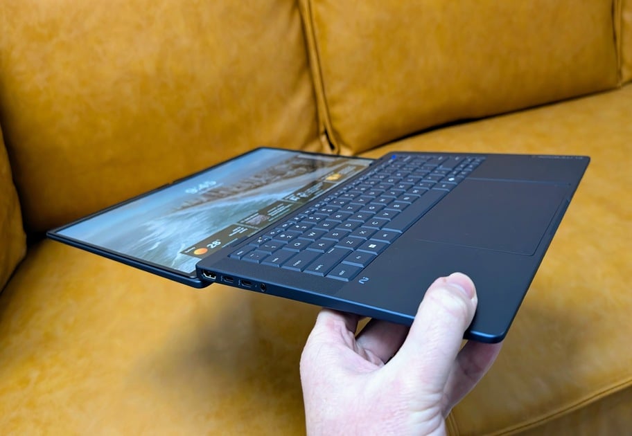 Hand holding a slim dark laptop partially opened, resting near a yellow sofa.